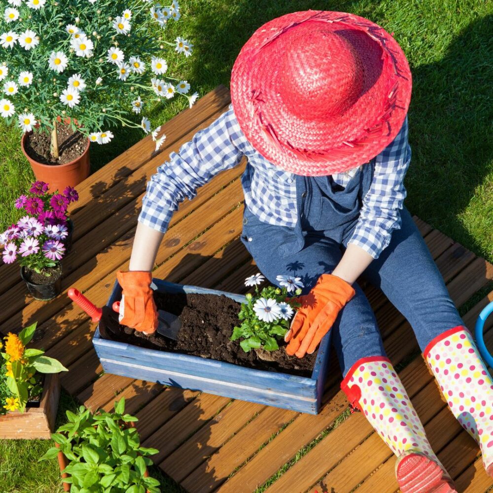 Image of person sitting in a garden. They're planting a white flower in a blue container. They are wearing a wide brimmed pink hat, blue overalls, a blue plaid shirt, and red polka dot boots.