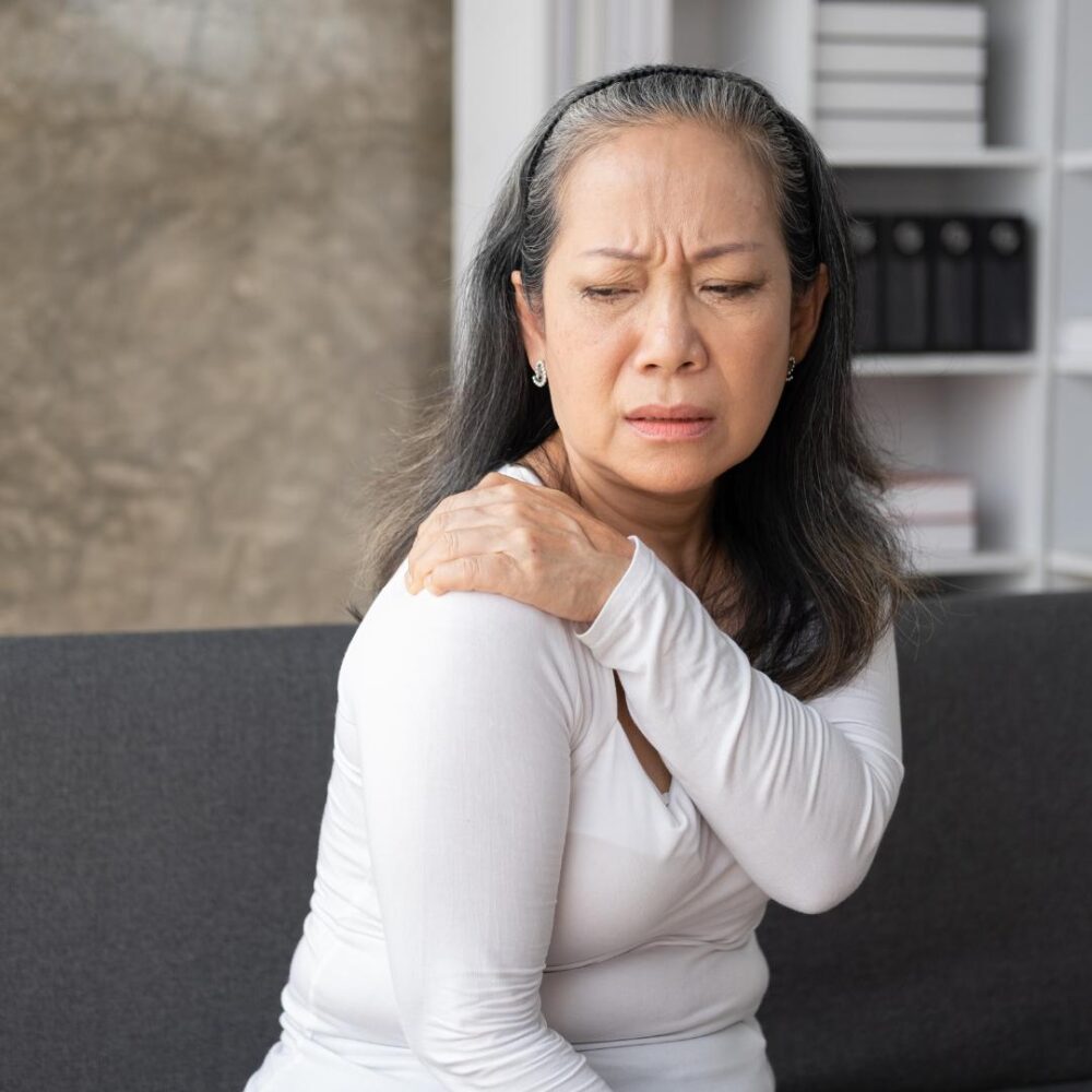 Photo of woman with long brown hair, wearing a white shirt. She's grabbing her shoulder in pain, as if she suffers from range of motion injuries.